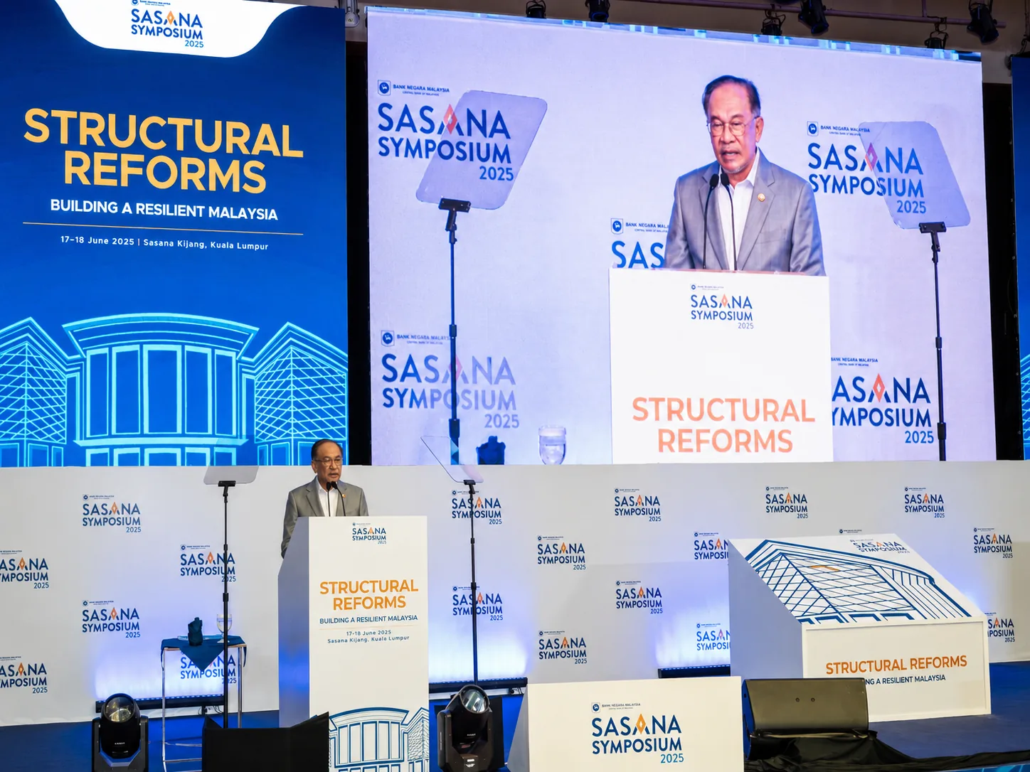 Sasana Symposium 2025 – keynote speaker at podium with large LED screen and side portrait screens